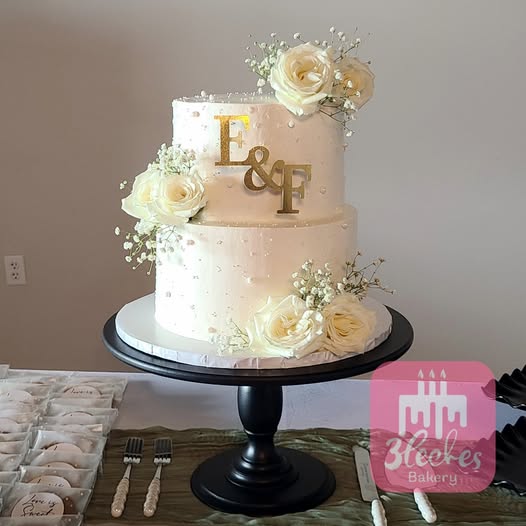 Elegant two-tier wedding cake with white buttercream, gold monogram, and fresh white roses